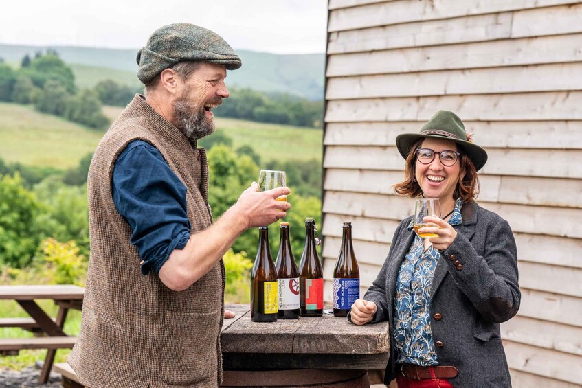 Two people holding glasses of beer, standing by a wooden table with bottles of beer in a rural outdoor setting. 1