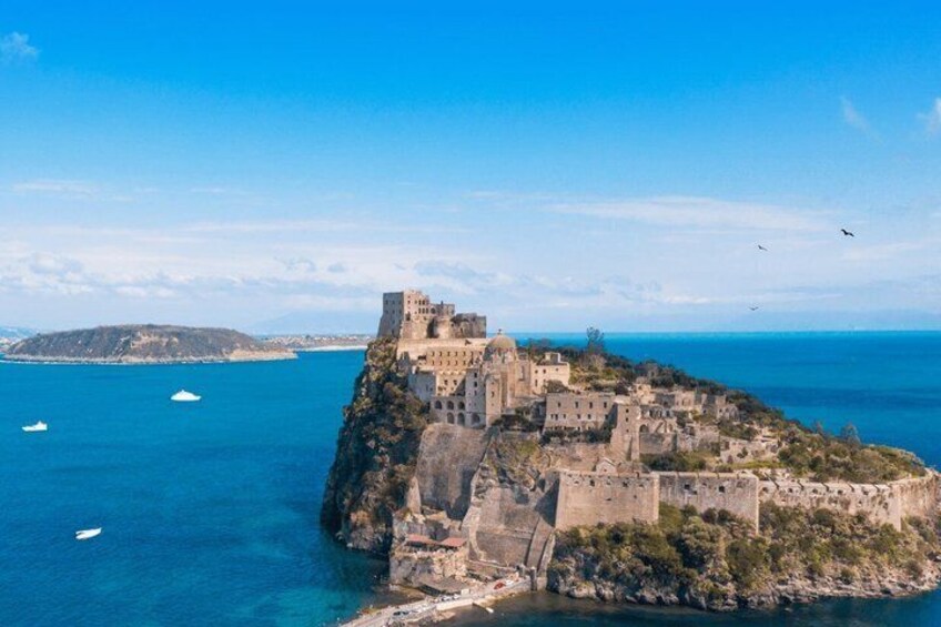 A coastal fortress with multiple buildings, a clear blue sea, and a distant island. 1