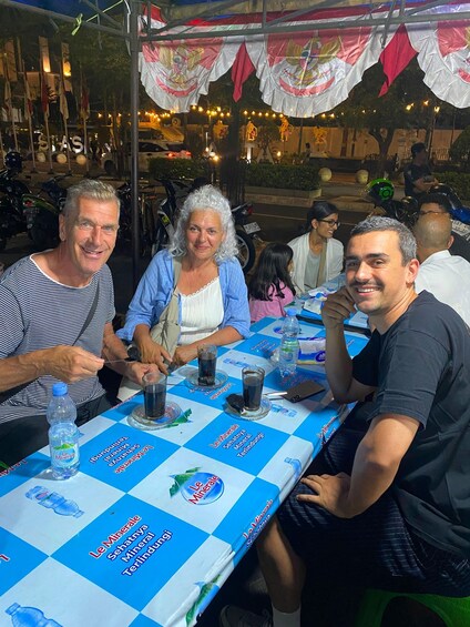A group of people seated at an outdoor table with a blue and white checkered tablecloth, under a canopy with hanging decorations. 2