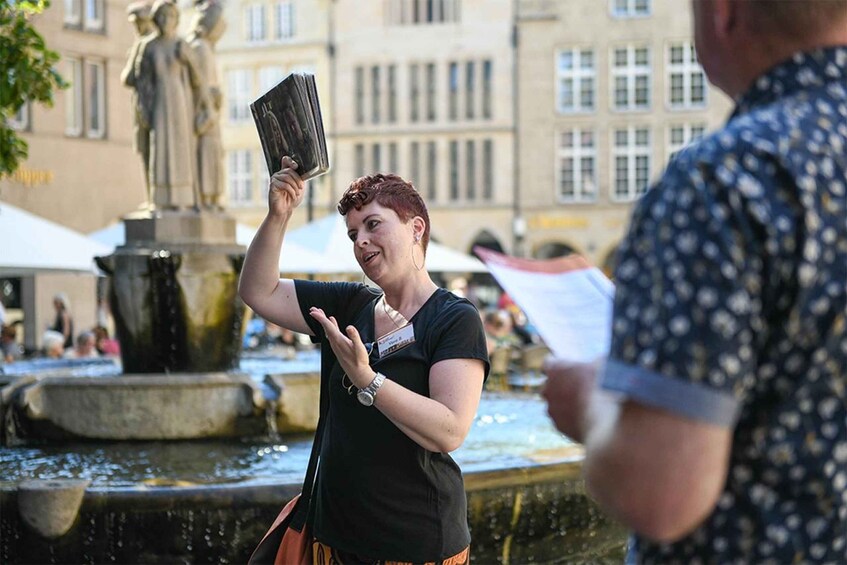 A person holding a book or tablet in an outdoor setting with a fountain and statues in the background. 2