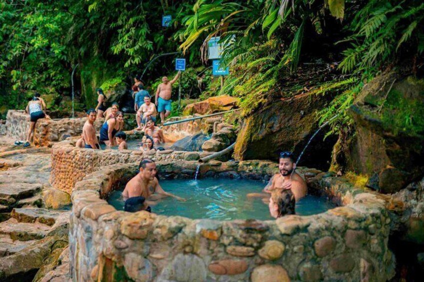 A natural hot spring with people bathing, surrounded by lush greenery and rocky terrain. 3