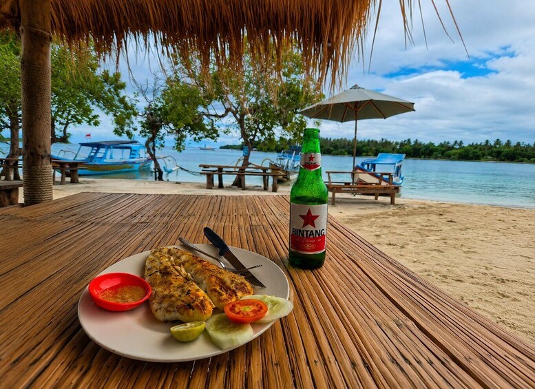 A wooden table with a plate of grilled chicken, a bottle of Bintang beer, and a red bowl of sauce on a beach with palm trees and boats in the background. 3