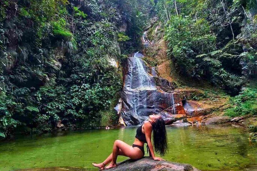 A waterfall, dense greenery, and a person sitting by a water body. 1