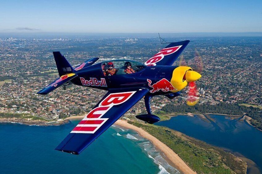 A Red Bull sponsored aircraft flying over a coastal landscape with a city in the background. 2