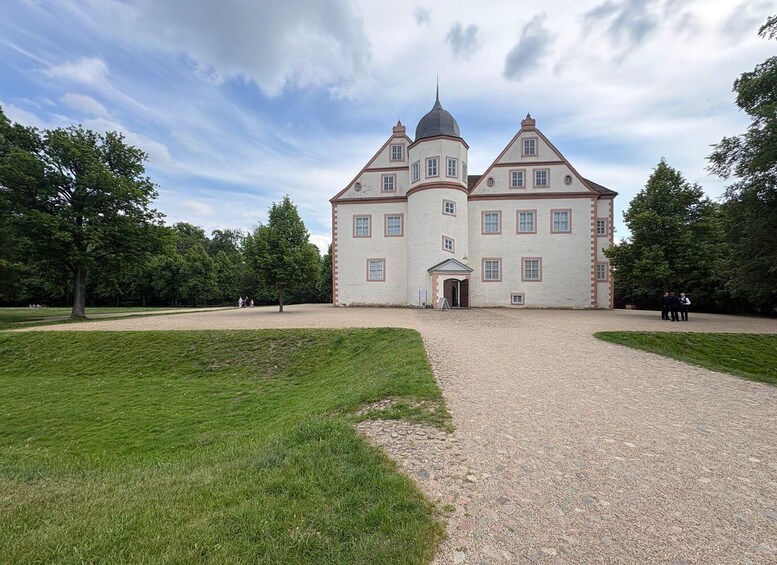 A large, historic building with a central dome and multiple windows, surrounded by a gravel driveway and lush green lawns. 1