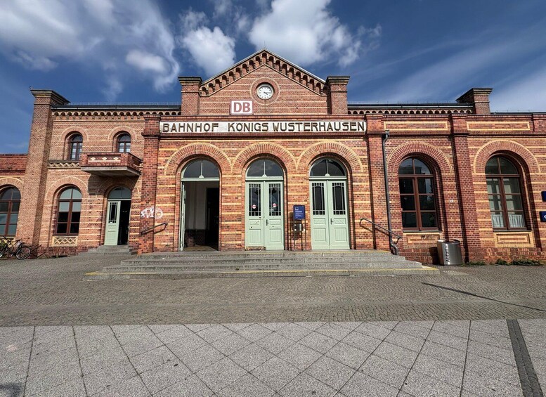 A historic building with arched windows and a clock, labeled 'BAHNHOF KONIGS WUSTERHAUSEN.' 4