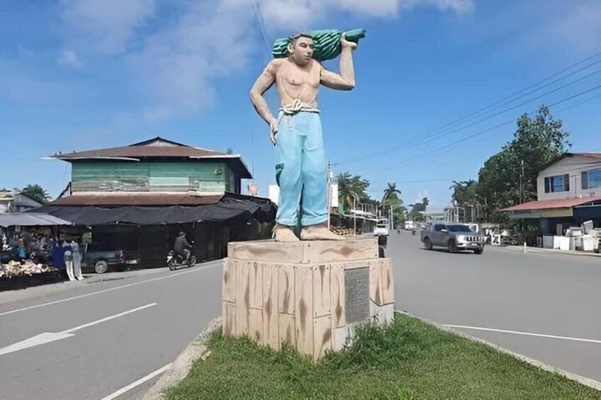 A statue of a man carrying a green object on a pedestal in a street with buildings and vehicles. 1