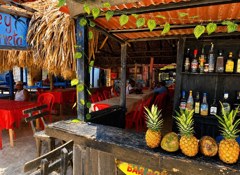 A rustic outdoor bar with a thatched roof, red plastic chairs, and a variety of bottles on shelves. 3