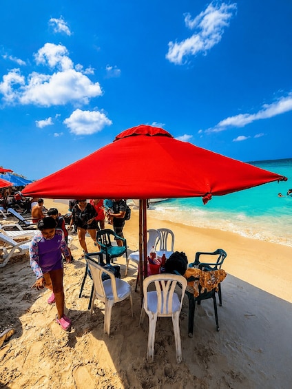 A beach with a red umbrella, white plastic chairs, and people walking. 4
