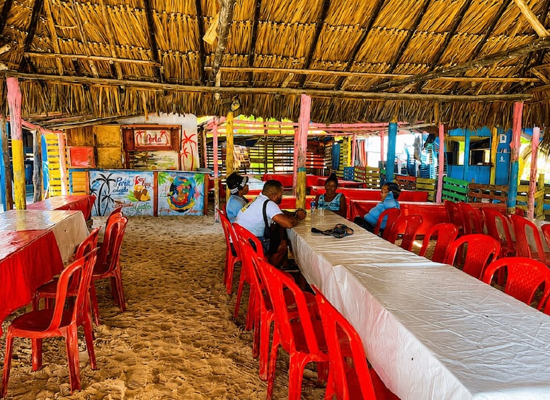 A thatched-roof dining area with red plastic chairs and tables, colorful murals, and a sandy floor. 5