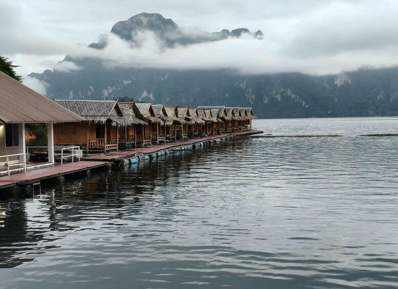 A row of thatched-roof overwater bungalows extending into a calm lake, surrounded by mountains and clouds. 5