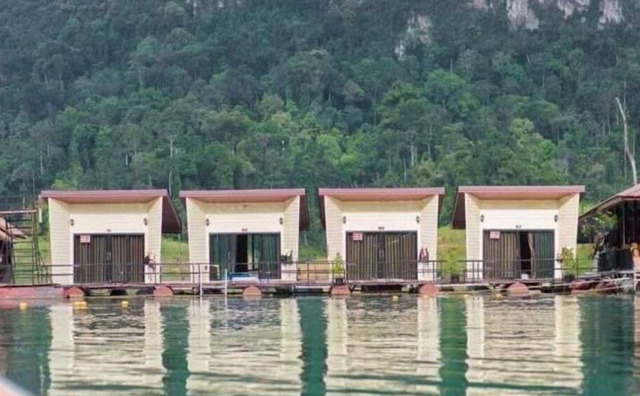 A row of small buildings with open doors, a body of water in the foreground, and a forested hillside in the background. 4