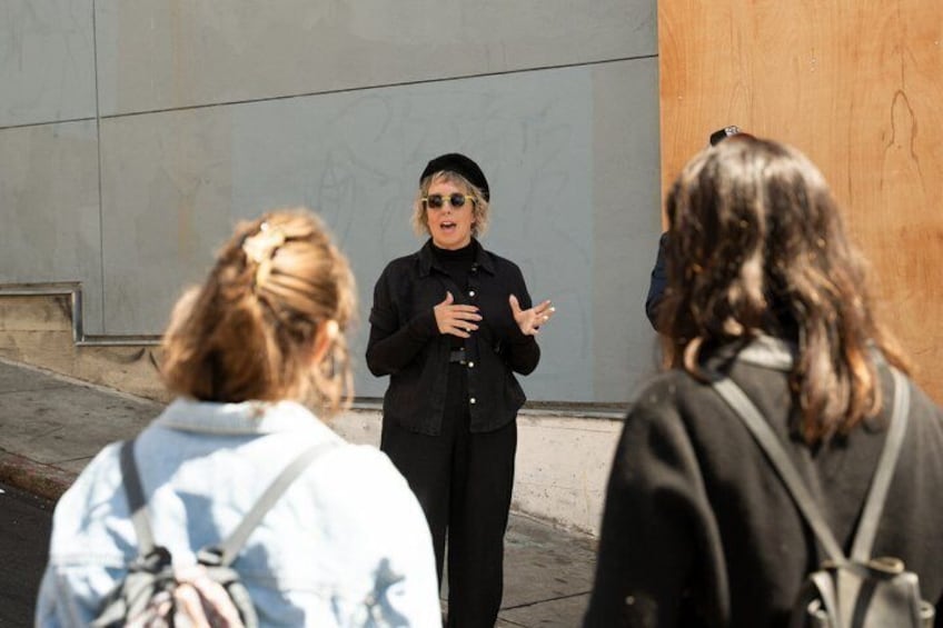 A person gesturing while speaking to two individuals in an urban setting with a concrete wall and a wooden structure in the background. 5