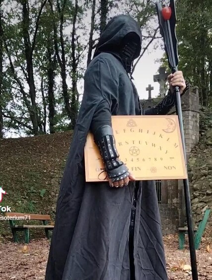 Une personne vêtue d’une cape à capuche, tenant un bâton et une carte ornée de symboles. 2