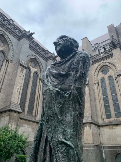 Une statue d’une femme vêtue d’une longue robe, se tenant devant un bâtiment aux fenêtres en arc et aux détails architecturaux travaillés. 5