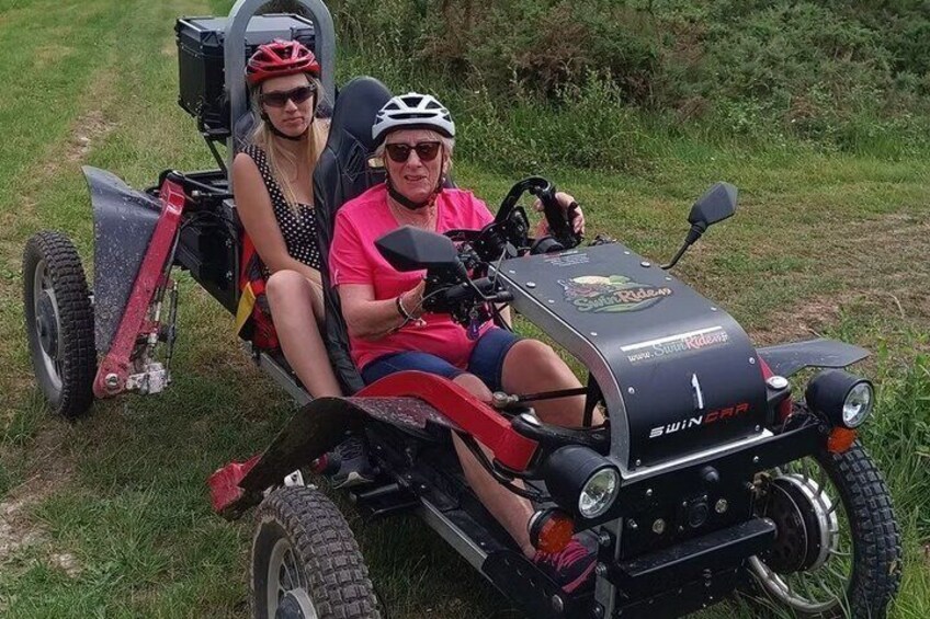 Two people in a red and black off-road vehicle with a trailer, on a grassy path. 4