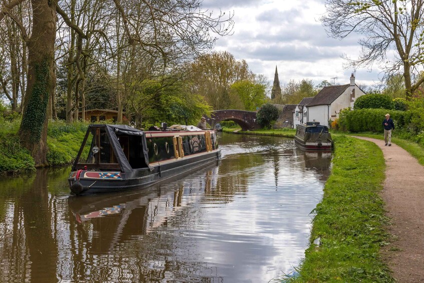 A narrow canal with a narrowboat docked on the left, a bridge in the background, and a white building with a chimney on the right. 1