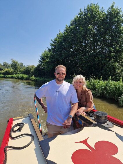 Two people on a narrowboat with a river and greenery in the background. 4