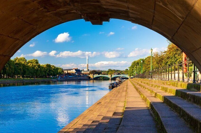A river under a stone bridge with steps leading down to the water, a distant tower, and a clear sky. 1