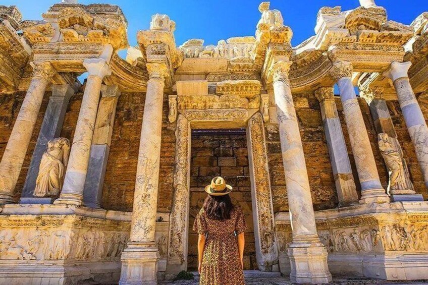 A person in a long dress and hat standing in front of a temple with columns and detailed carvings. 1