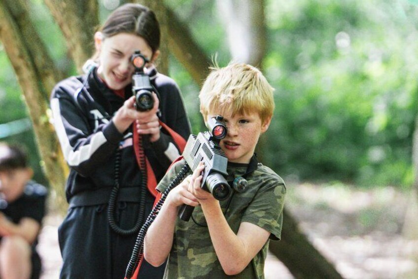 A person in camouflage holding a firearm with a scope, another person in a black jacket with a camera, and a forested background. 5