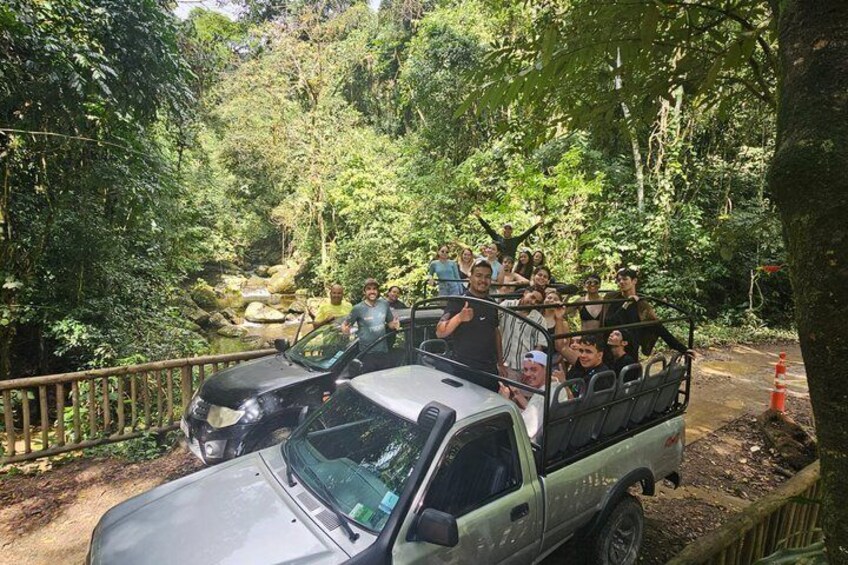 A group of people on a safari vehicle in a forested area with a river and a bridge. 1