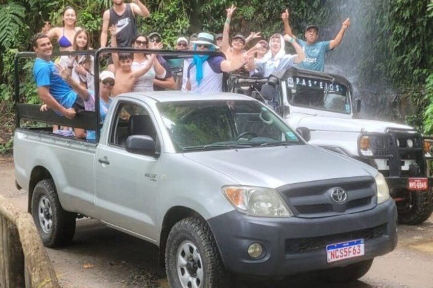 A silver pickup truck with a group of people standing on the back, some raising their hands. 4
