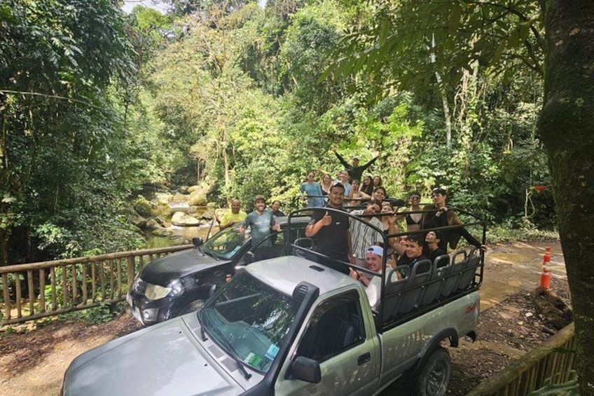 A group of people on a safari vehicle surrounded by dense forest and a small stream. 1