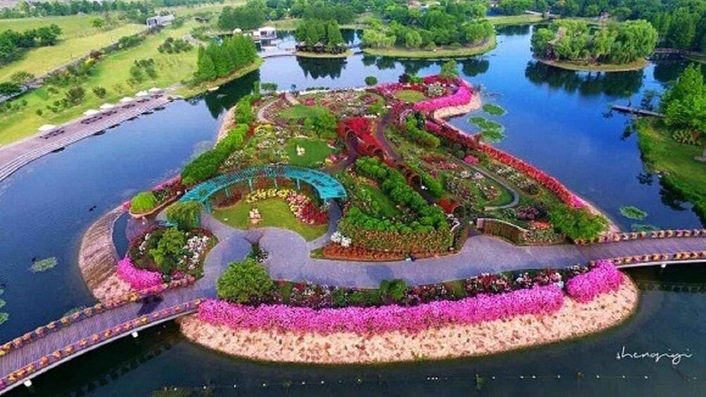 An aerial view of a landscaped island with a winding road, flower beds, and a bridge. 1