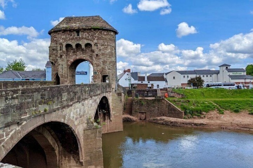 A stone bridge with a tower, a river, and buildings in the background. 4