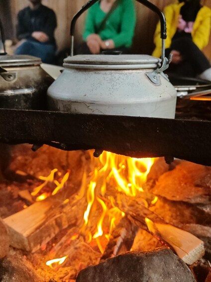 A close-up of a white kettle on a metal stand above a campfire with logs and flames. 2