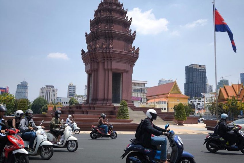 A busy street with motorbikes and scooters, a large red monument, and modern buildings in the background. 5