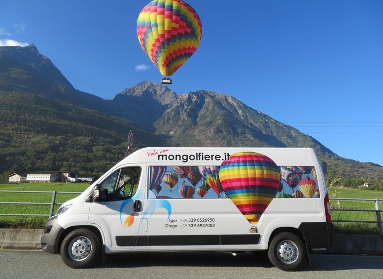 A white van with colorful hot air balloons on the side, parked on a road with a mountainous landscape in the background. 4