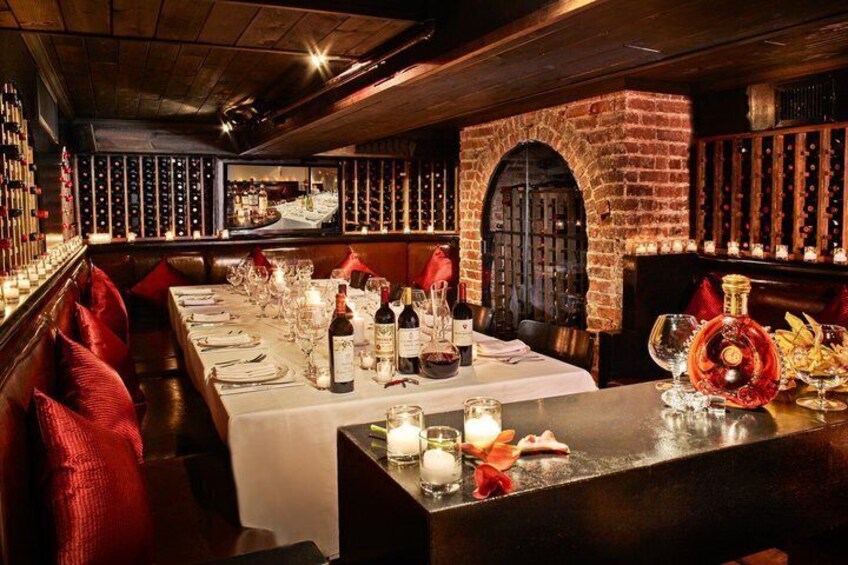 A restaurant interior with a long dining table set for a meal, a brick fireplace, and wine bottles. 4