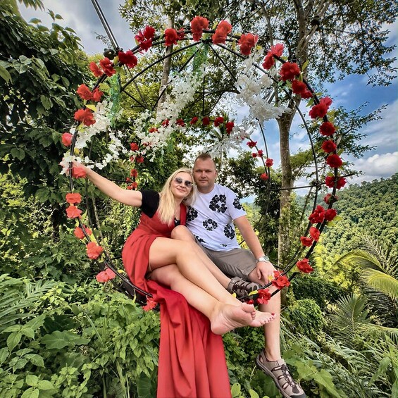 A couple sitting on a swing decorated with red and white flowers, surrounded by lush greenery. 1