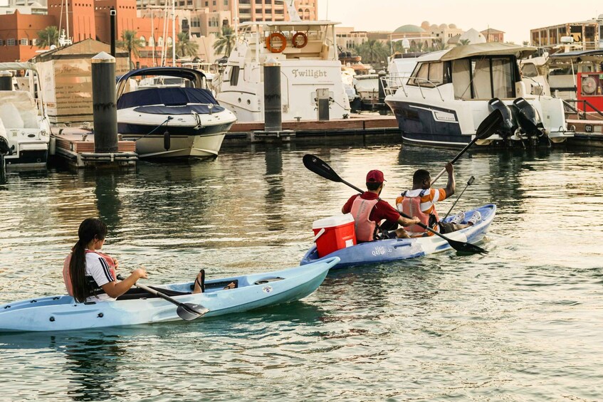 Three people rowing a light blue canoe on calm water, with boats docked in the background. 3