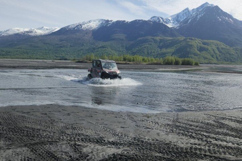 A vehicle driving through a shallow river with mountains in the background. 5