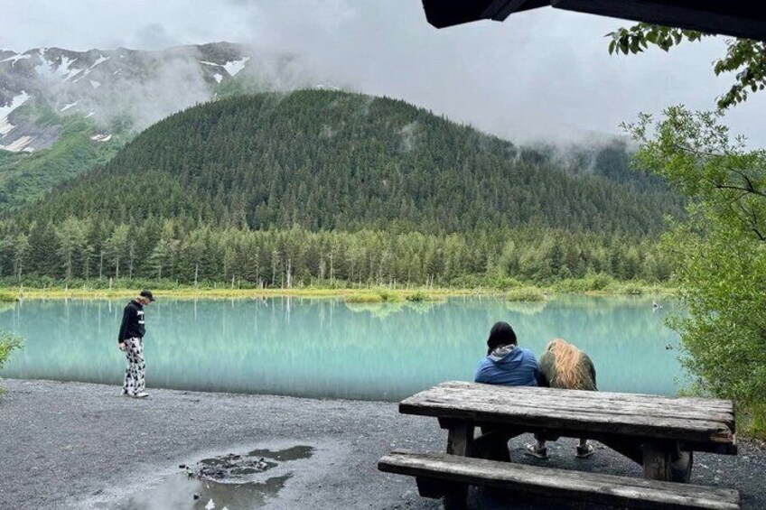 A lake with a wooden bench, a person standing by the water, and a forested mountain in the background. 2