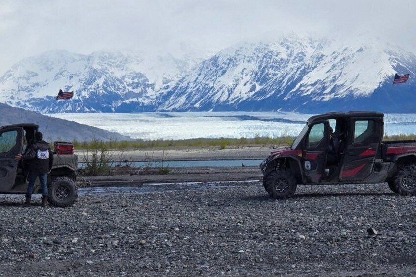 Two off-road vehicles on a gravel path with a glacier and mountains in the background. 1