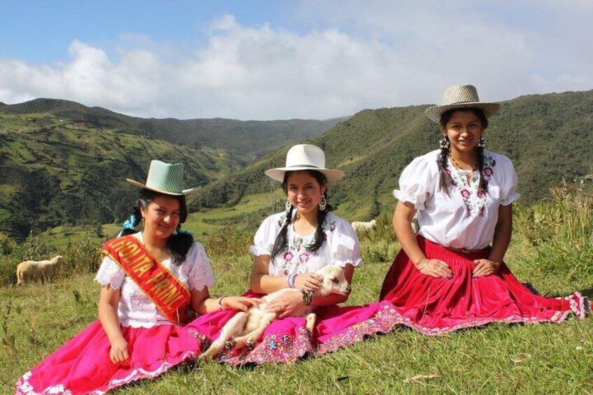 Three women in traditional attire sitting on grass with a mountainous landscape in the background. 1