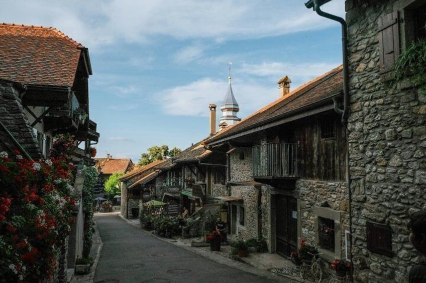 A narrow street lined with traditional stone and timber buildings, a church steeple in the distance, and flowering plants along the sidewalk. 5