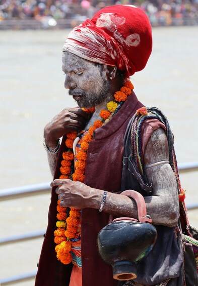 A person wearing a red turban and a maroon garment, adorned with orange flowers, standing by a body of water. 5
