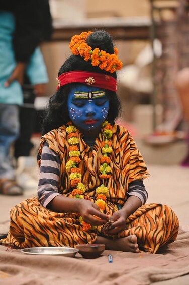 A person sitting on the ground, wearing a tiger-striped outfit and a red headband, with a garland of marigold flowers around their neck. 1
