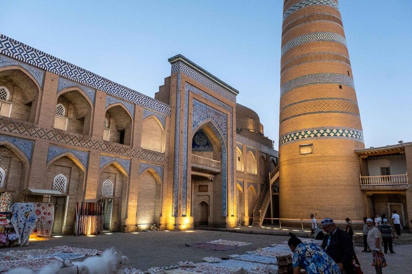 A historic building with intricate blue and white tile work, a large central tower, and a courtyard with people and traditional rugs. 4