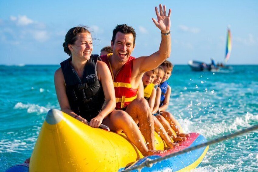A group of people on a yellow and blue raft in the ocean with a sailboat in the background. 5
