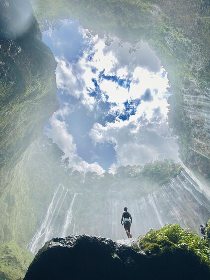 A waterfall cascading down a cliff, a person standing on a rock, and a clear sky above. 2