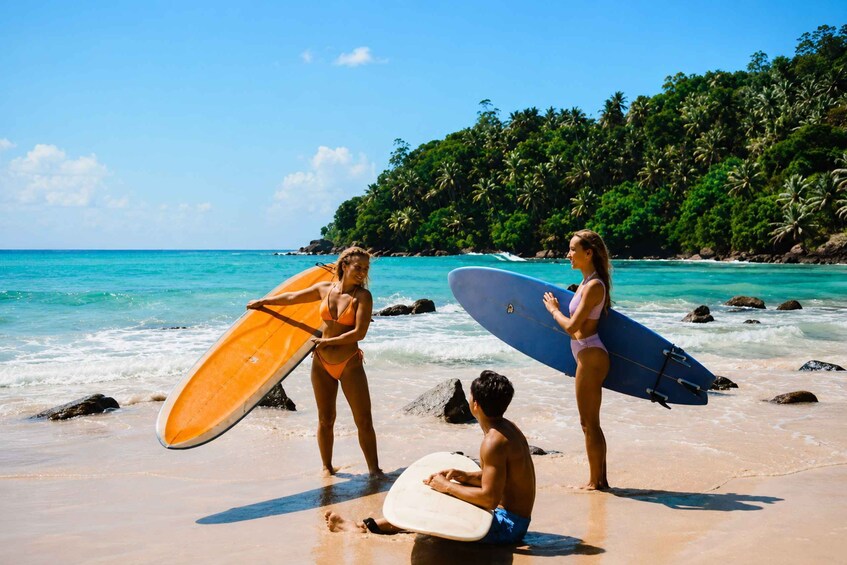 Two people on a beach holding surfboards, with clear turquoise water and a forested hill in the background. 3