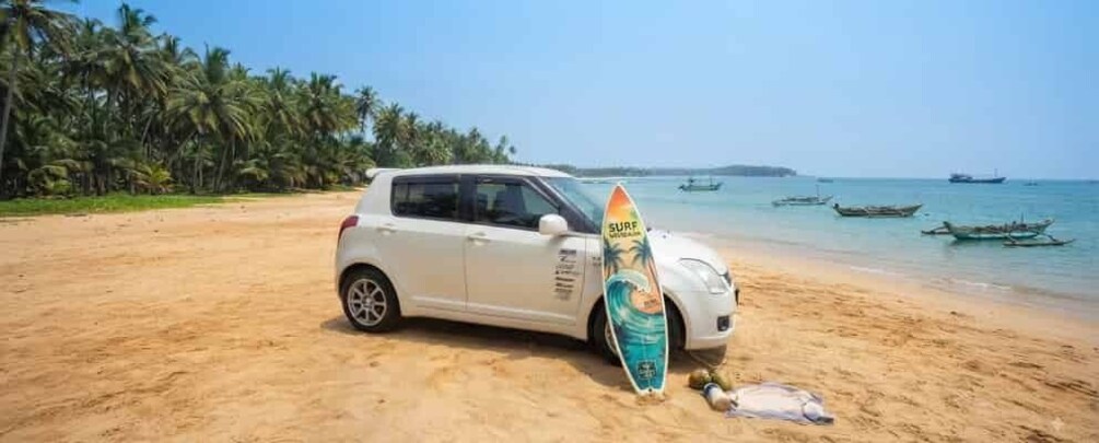 A white car parked on a sandy beach with a surfboard leaning against it. 1