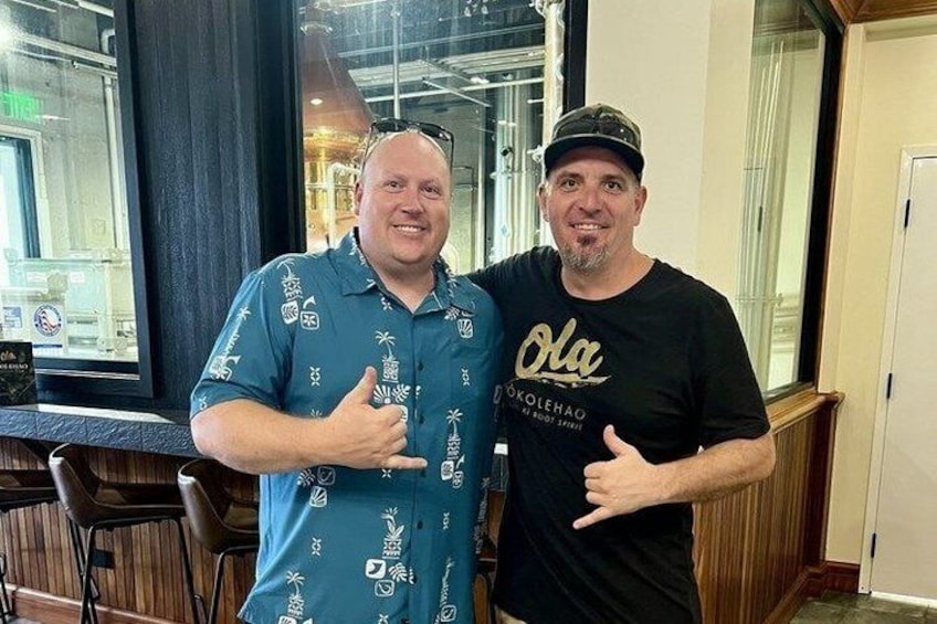 Two men standing indoors, one wearing a blue shirt with patterns and the other in a black shirt with text. 3