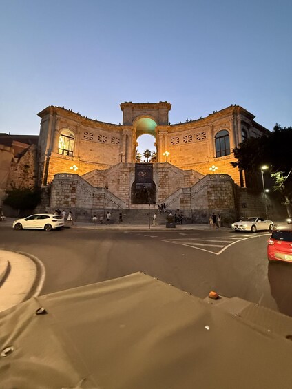 A historic stone archway with illuminated windows and a central arch, surrounded by a road with cars and pedestrians. 3